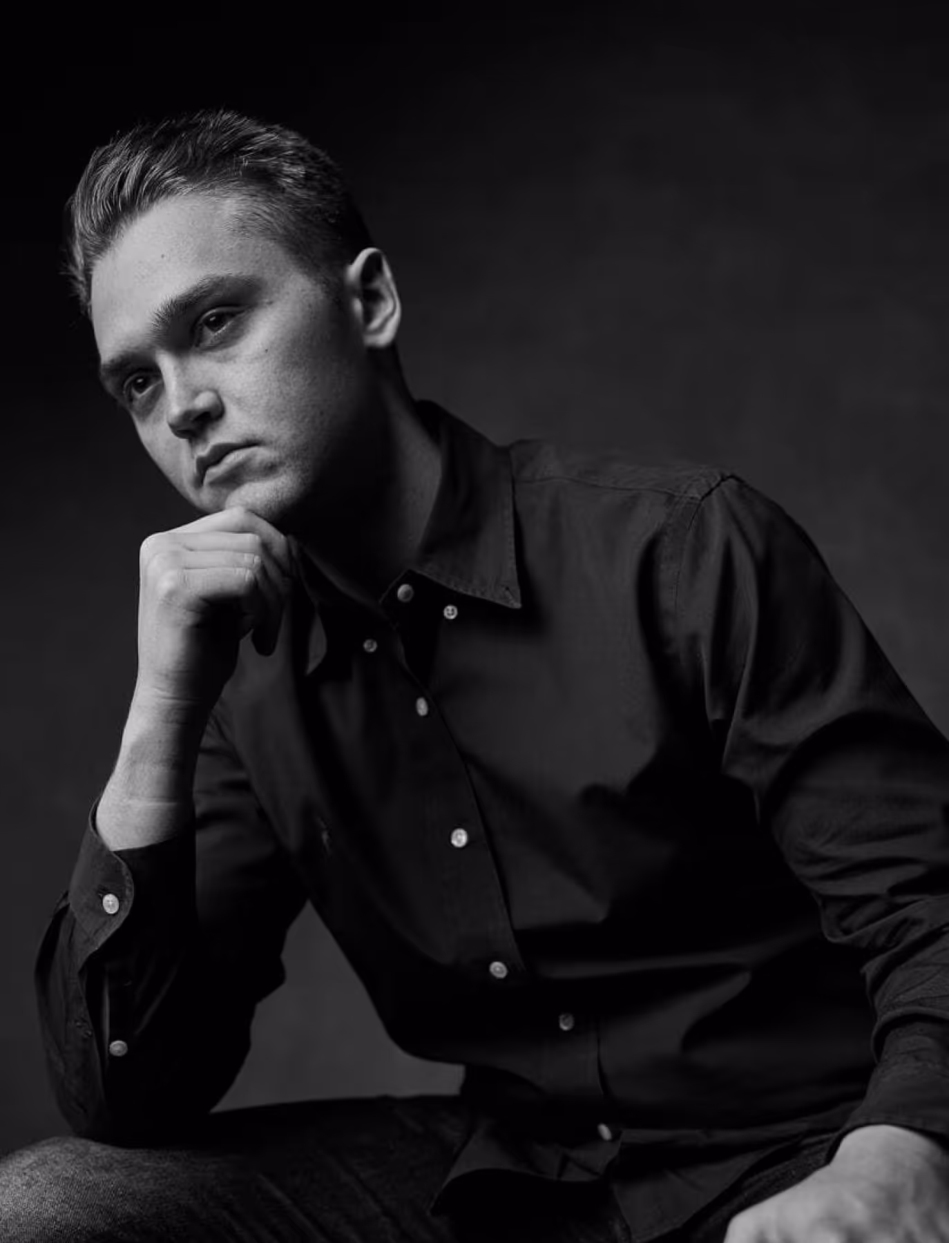 Black and white portrait of a young man in a dark button-up shirt resting his chin on his hand, looking thoughtful.